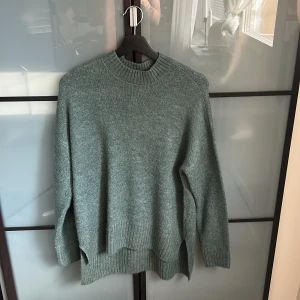 Grön stickad tröja från Vero Moda - Mysig grön stickad tröja från Vero Moda med rund halsringning och ribbade muddar. Tröjan har en loose passform och slitsar i sidorna, perfekt för lager på lager under kyliga dagar.