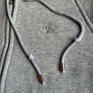 Grå hoodie från Burberry med rutig huva - Grå hoodie från Burberry med klassiskt rutigt foder i luvan och insidan. Tröjan har dragkedja, snörning i huvan och broderad logga på bröstet. Perfekt för dig som vill ha en stilren och lyxig look med ikoniska Burberry-detaljer. För flera bilder eller frågor så är de bara att slå ett pling sick 10/10 priset kan diskutera vid snabb affär. 