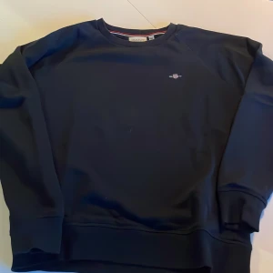 Mörkblå sweatshirt från GANT - Säljer en stilren mörkblå sweatshirt från GANT med broderad logga på bröstet. Tröjan har rund halsringning med ribbade muddar och en snygg detalj med röd och vit rand på insidan av kragen. Perfekt för en avslappnad look. Storlek 158-164