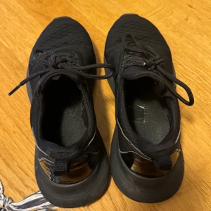 Svarta sneakers med snörning - Säljer ett par helsvarta sneakers med sportig look och platt sula. Ovandelen är i textil med diskret mönster och snörning framtill. Skorna har dragflik bak och detaljer i syntet vid hälen. Perfekta för dig som gillar stilrena och bekväma skor.