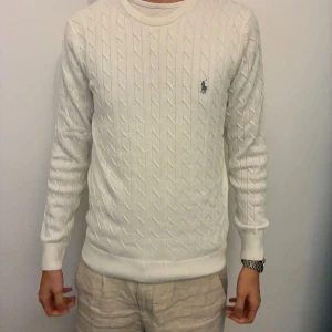 Vit kabelstickad tröja från Ralph Lauren - Vit kabelstickad tröja från Ralph Lauren med klassisk rund halsringning och broderad logga på bröstet. Tröjan har lång ärm och ribbade muddar vid ärmslut och nederkant. Perfekt för en clean och stilren look.