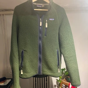 Grön fleecejacka från Patagonia M - Mysig grön fleecejacka från Patagonia med hel dragkedja framtill och tre fickor med dragkedja. Jackan har en schysst struktur och står ut med sin mörkgröna färg och blå detaljer vid dragkedjorna. Perfekt för lager-på-lager under kyliga dagar.