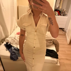 Beige skjortklänning med knappar - Snygg beige skjortklänning med korta ärmar, krage och guldiga knappar hela vägen framtill. Klänningen har två bröstfickor och markerade sömmar som ger en figurnära passform. Perfekt för dig som gillar en stilren och trendig look. helt ny, prislapp kvar 