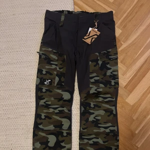 RVRC camo cargopants med fickor - Snygga cargopants från RVRC med mörkgrå bas och camouflage-mönstrade paneler. Byxorna har flera praktiska fickor, förstärkta knän och elastisk midja med bälteshällor. Oanvända med lappen kvar 