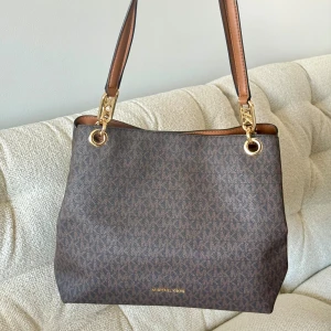 Brun axelväska från Michael Kors - Nypris: 4.400 kr             Snygg brun axelväska från Michael Kors med MK-mönster över hela väskan. Väskan har guldfärgade detaljer, bruna handtag och en praktisk dragkedja upptill. Materialet är skinn och syntet, och insidan har flera fack för dina grejer. Perfekt storlek för att få plats med allt du behöver.