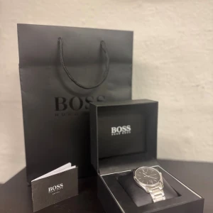 Silverfärgad klocka från Hugo Boss - Stilren klocka från Hugo Boss med svart urtavla och silverfärgad länk i metall. Klockan har datumvisning. Perfekt accessoar för dig som gillar klassisk och modern stil. Nypris cirka 4500. Klockan har en liten repning, går att fixa enkelt.