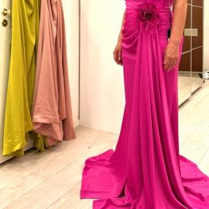 Cerise långklänning med blomdetalj - Superfin cerise långklänning från MNM Couture i glansigt tyg. Klänningen har draperad midja, stor blomapplikation med fjädrar på höften och en elegant slits framtill. Sitter figurnära och har ett långt släp för extra drama.