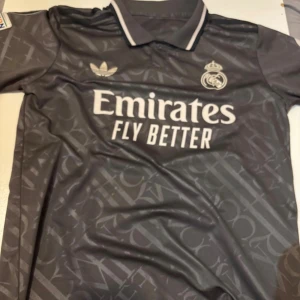 Real Madrid Bellingham fotbollströja - Säljer en mörkgrå Real Madrid fotbollströja från Adidas med Bellingham och nummer 5 på ryggen. Tröjan har korta ärmar, broderat klubbmärke, klassisk krage och Champions League-märke på ärmen. Materialet är lätt och andas, perfekt för match eller träning.