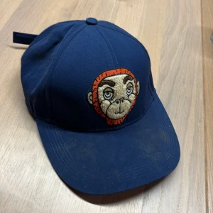 Mini Rodini Blå keps med broderad apa - Mini Rodini keps med justerbar rem och broderad apa framtill i orange och beige. Klassisk form med böjd skärm och ventilerande hål. Fint skick men behöver tvättas stl 52/54