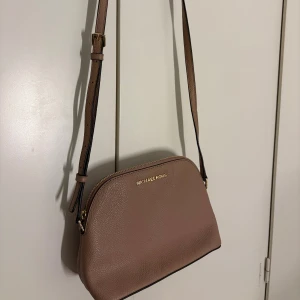 Beige/ljusrosa axelväska från Michael Kors - Snygg beige axelväska från Michael Kors i stilren design. Väskan är tillverkad i strukturerat skinn och har justerbar axelrem samt guldfärgad logga framtill. Perfekt storlek för det viktigaste, passar till många outfits och även som en daglig väska. 