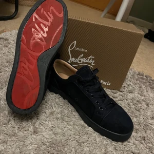 Christian Louboutin blåa sneakers mocka - Snygga svarta sneakers från Christian Louboutin i mocka med klassisk röd sula och rund tå. Skorna har svarta snören, diskret logga bak och en stilren design som passar till det mesta. Insidan är ljusbeige och sulan är platt för en clean look.