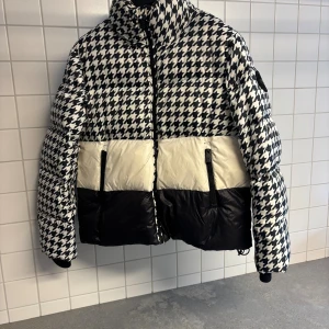 Svartvit pufferjacka från 8848 Altitude - Snygg pufferjacka från 8848 Altitude med svartvitt hundtandsmönster upptill, vit mittsektion och svart nedtill. Jackan har hög krage, dragkedja framtill och två fickor med dragkedja. Yttermaterialet lätt och vindtätt, fyllning premium dun med 650 fyllnadsgrad, extra kraftiga dragkedjor från YKK.. På insidans nederdel sitter ett brett, ribbstickat snölåsmed tryckknappar och i ärmsluten finns integrerade muddar för att stänga snö och kyla ute. 