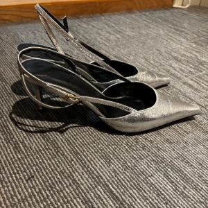 Klackar - (Använda ENDAST en gång!) Säljer ett par glansiga silvriga pumps med smal klack och spetsig tå. Skorna har öppen häl och smala remmar med spänne för en snygg och elegant look. As snygga och trendiga från H&M. 