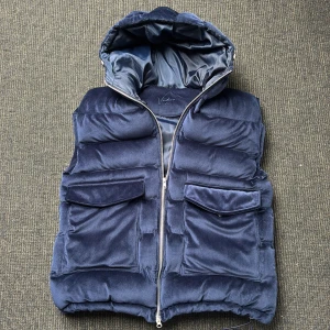 Mörkblå manchester dunväst  - Snygg mörkblå dunväst i mjuk manchester. Västen har en vadderad look, stor huva, två stora fickor framtill och dragkedja hela vägen. Perfekt för lager-på-lager och ger en cool vibe till din outfit. Saknar 1 knapp för den högra fickan. Anars bra skick i storlek L.