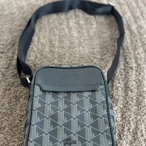 Svart axelväska från Lacoste - Snygg svart axelväska från Lacoste med geometriskt mönster i vitt. Väskan har justerbar rem, dragkedja och detaljer i skinnimitation. Perfekt storlek för att bära med sig det viktigaste och har en modern look med tydlig Lacoste-logga, Köpt från själva Lacoste affären