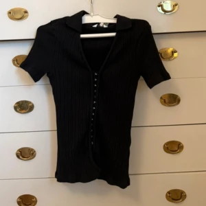 Svart ribbad kortärmad topp med knappar - Svart ribbad topp med korta ärmar och v-ringning. Toppen har en snygg krage och knappar framtill som ger en cool detalj. Modellen är croppad och sitter tajt mot kroppen, perfekt till jeans eller kjol.