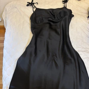 Svart långklänning från NA-KD Reborn - Säljer en svart silkes klänning med längden till knäna från NA-KD Reborn med smala axelband som knyts på axlarna. Klänningen har en slät, glansig yta och en figurnära passform med lätt rynk vid bysten. Perfekt för dig som gillar stilrena och eleganta plagg.