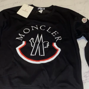 Svart Moncler sweatshirt med logga - Svart sweatshirt från Moncler med stor vit och röd logga framtill. Tröjan har rund halsringning, långa ärmar och ribbade muddar. Klassisk och stilren design med Moncler-märke på ärmen. Perfekt för dig som gillar streetwear och premiumkänsla.