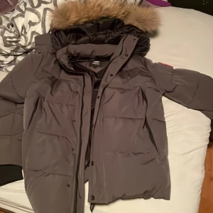 Grå dunjacka med fuskpäls - Säljer en grå dunjacka från Didriksons med stor huva och fuskpälsdetalj. Jackan har dragkedja och knappar framtill, stora fickor och är perfekt för kalla dagar. Klassisk och stilren design som håller dig varm hela vintern.