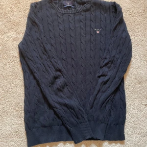 Mörkblå kabelstickad tröja från GANT - Kabelstickad mörkblå tröja från GANT i 100% bomull. Klassisk rund halsringning, ribbade muddar och broderad GANT-logga på bröstet. Perfekt för dig som gillar en tidlös och stilren look.