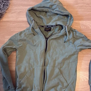 Grön hoodiejacka från Denim Day M - Grön hoodiejacka från Denim Day med dragkedja framtill, snörning i huvan och två fickor. Jackan har en svart patch på ena ärmen och är tillverkad i mjuk bomull. Perfekt för chill dagar och enkel att matcha med jeans eller joggers.