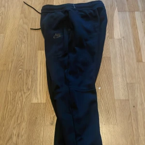 Svarta mjukisbyxor från Nike, stl L (147-158cm) - Svarta mjukisbyxor från Nike i storlek L ( 147-158cm) med dragsko i midjan och diskret Nike-logga på benet. Byxorna har en avslappnad passform och är tillverkade i mjukt material, perfekta för chill eller träning. Klassisk look med fickor och elastisk midja.