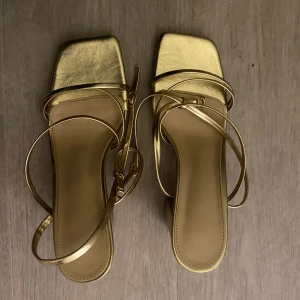 Guldiga sandaler med klack och remmar - Säljer ett par trendiga sandaler i metallic guld med fyrkantig tå och smala remmar som går runt foten. Skorna har blockklack och öppen tå, vilket ger en modern och stilren look. Perfekta för dig som vill sticka ut med en glansig detalj till outfiten. 39/40