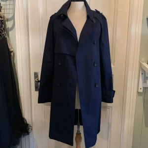 Mörkblå trenchcoat med bälte - Snygg mörkblå trenchcoat med klassisk dubbelknäppning, axelklaffar och bälte i midjan. Jackan har långa ärmar med spännen vid handlederna och en stilren krage. Perfekt för dig som vill ha en tidlös och clean look.