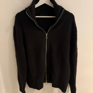 Stickad fullzip tröja - Mysig svart stickad kofta från Brand Island med hög krage och hel dragkedja framtill. Perfekt för lager-på-lager och har ribbade detaljer vid krage och muddar. Enkel och stilren design som funkar till allt.