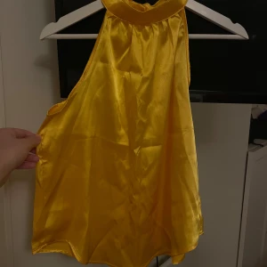 Gul halterneck topp från SHEIN - Säljer en snygg gul halterneck topp från SHEIN i glansigt satinliknande material. Toppen är ärmlös med hög krage och har en lös passform som ger en cool och trendig vibe. Perfekt för att sticka ut med färg och stil.