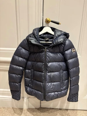 Mörkblå dunjacka från Colmar - Snygg mörkblå pufferjacka från Colmar med huva och quiltade detaljer. Jackan har dragkedja framtill, två sidofickor och ett diskret Colmar-märke på ärmen. Perfekt för kalla dagar och ger en sportig vibe. Jackan strl motsvarar 34