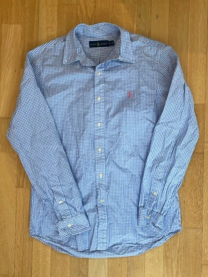 Blårutig skjorta från Ralph Lauren - Klassisk blårutig skjorta från Ralph Lauren i 100% bomull. Skjortan har lång ärm, knappar framtill och den ikoniska röda loggan broderad på bröstet. Perfekt till jeans eller chinos för en clean och tidlös look.
