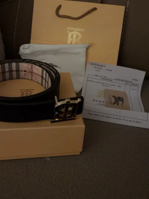 Rutig Burberry bälte med B-spänne - Säljer ett klassiskt Burberry bälte med det ikoniska rutiga mönstret i beige, brun och röd. Bältet har ett stort metallspänne med Burberrys B-logga och insida i svart. Kommer med originalpåse och box. De passar för olika stilar och kan bli i både svart färg och beige! Man kan göra extra hål enkelt om den var stor