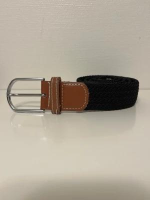 Svart flätad bälte med läderdetaljer - Snyggt svart flätat bälte med bruna läderdetaljer och silvrig spänne.