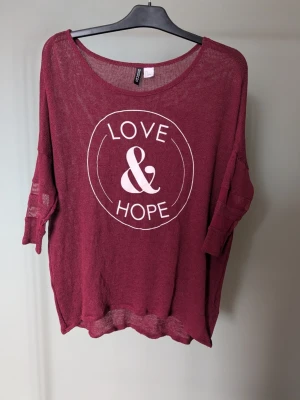 Vinröd meshtröja från Divided - Säljer en vinröd meshtröja från Divided H&M med trycket 'LOVE & HOPE' i vitt framtill. Tröjan har lös passform, rund halsringning och trekvartsärmar med dekorativa banddetaljer. Materialet är tunt och genomskinligt, perfekt för lager på lager.