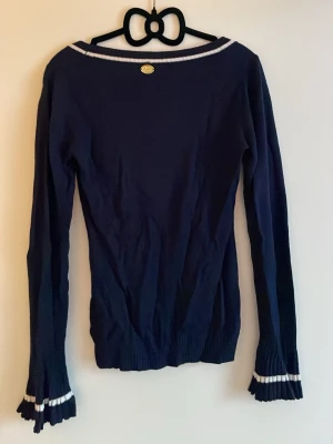 Marinblå långärmad topp med volang på ärmar - Säljer en marinblå långärmad topp från H&M med vita ribbade detaljer vid halsringning, ärmslut och dekorativa fickor framtill. Toppen har volangkant på ärmarna och guldknappar på fickorna för en extra touch. 