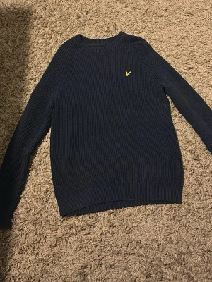Mörkblå stickad tröja Lyle & Scott - Säljer en mörkblå stickad tröja från Lyle & Scott med klassisk gul logga på bröstet. Tröjan har ribbade muddar, rund hals och lång ärm. Perfekt för dig som gillar stilrena och tidlösa plagg.