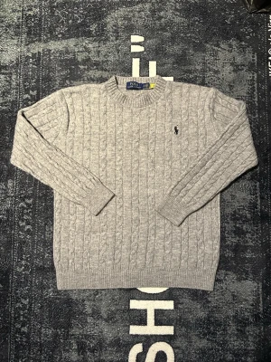 Grå kabelstickad tröja Polo Ralph Lauren - Kabelstickad grå tröja från Polo Ralph Lauren med klassisk rund hals och broderad logga på bröstet. Tröjan är långärmad och har ribbade muddar vid ärmslut och nederkant. Perfekt för lager på lager under kyliga dagar.
