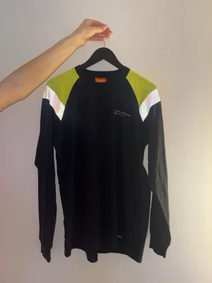 Svart långärmad arbetströja Tranemo - Svart långärmad arbetströja från Tranemo med gula axelpartier och reflekterande detaljer. Tryck på bröstet och rygge. Tillverkad i flamskyddat material.