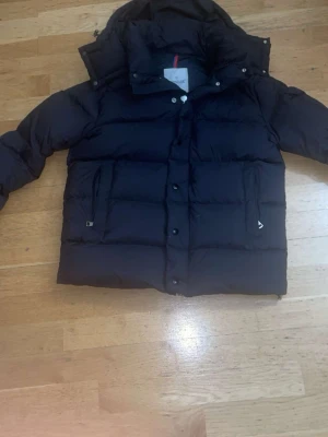 Svart Moncler Vezere Jacka - Säljer en svart vezere från Moncler med huva och klassisk logga på ärmen. Jackan har dragkedja och knappar framtill, två fickor med dragkedja och ett coolt innerfoder med serietidningsmönster. Perfekt för kalla dagar och riktigt snygg. Pris diskuteras pm 
