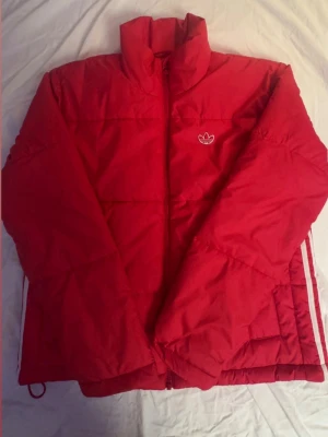 Röd pufferjacka från Adidas - Säljer en röd pufferjacka från Adidas med klassisk Trefoil-logga broderad på bröstet. Jackan har hög krage, dragkedja framtill och diskreta vita ränder på sidan. Perfekt för kalla dagar och riktigt snygg streetstil.