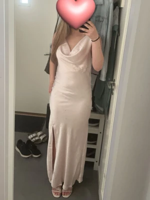 Ljusrosa balklänning - Säljer en elegant balklänning i ljus champagnefärg med slits framtill och draperad urringning. Smutsen som syns på bilden är från spegeln så det är ingen skada på klänningen. Ryggen är v-ringad ner till en bit mellan skulderbladen. Klänningen är ärmlös och är mjuk i satin tyg. Perfekt för dig som snart har bal. Lappen sitter kvar på ryggen, klänningen är använd en gång och är som i nyskick. Den är köpt från raglady by tara. Priset kan diskuteras.
