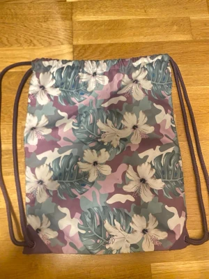 Blommig ryggsäck med snörning - Säljer en snygg gympåse/ryggsäck med blommigt mönster i lila, grönt och vitt. Väskan har snörning och är perfekt att slänga över axeln när du ska iväg. Materialet känns lätt och smidigt, och blommorna ger en fräsch vibe.