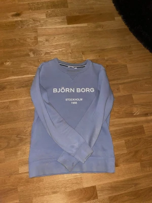 Ljusblå sweatshirt från Björn Borg - Säljer en ljusblå sweatshirt från Björn Borg med tryck på bröstet där det står 'BJÖRN BORG STOCKHOLM 1956'. Tröjan har rund halsringning, långa ärmar och är gjord i mjuk bomull. Perfekt för chill dagar eller när du vill ha en enkel och clean look. OBS! Fläckar förekommer på ena armen och längst ner på tröjan, vet ej vart det kommer från annars perfekt. 