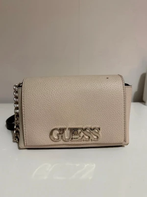 Beige axelväska från Guess - beige axelväska från Guess med guldfärgad logga framtill. Väskan har justerbar svart axelrem i skinnimitation och kedjedetaljer. Insidan är svart och har ett fack. Det finns en skada på väskan (bild 4) utöver det är den i bra skick.