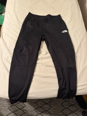 The North Face - Säljer ett par svarta mjukisbyxor från The North Face med logga på både fram- och baksidan. Byxorna har resår i midjan, smal passform och vita detaljer längs benen. Perfekta för chill eller träning och riktigt sköna att ha på sig.