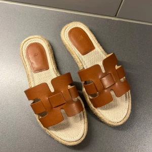 Bruna sandaler från H&M i storlek 36 - Säljer ett par snygga sandaler från H&M i storlek 36. De har breda bruna remmar i skinnimitation och en beige sula med flätad kant i jute. Perfekta för sommaren och riktigt trendiga till jeans eller klänning.