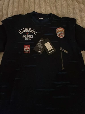 Svart DSQUARED2 t-shirt med patchar - Svart t-shirt från DSQUARED2 med broderade patchar och tryck på bröstet, inklusive texten 'BROMANCE' och 'CANADIAN TWINS'. Snygg dragkedjedetalj på framsidan. Klassisk rund hals och korta ärmar. Perfekt för dig som gillar streetstyle.