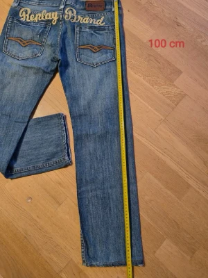 Blå Replay Brand jeans med broderi - Säljer ett par klassiska blå jeans från Replay Brand med snyggt broderad logga på bakfickorna och kontrastdetaljer. Jeansen har rak passform, normal midja och är tillverkade i slitstark bomullsdenim. Perfekta för dig som gillar en tidlös och avslappnad look. Dem är i bra skick inga skador.
