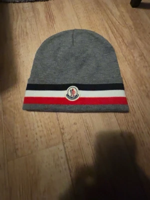 Grå Moncler mössa med ränder - Snygg grå mössa från Moncler med klassiska ränder i svart, vitt och rött längst ner. Mössan har en Moncler-logga framtill och är tillverkad i mjukt material som känns skönt mot huvudet. Perfekt accessoar för att lyfta din vinterstil.